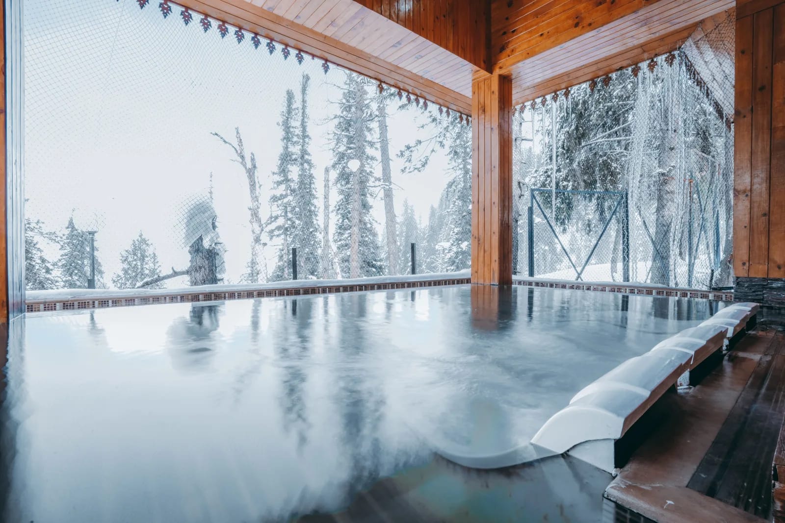 Steaming open-air bath under a timber roof with snowy conifers beyond