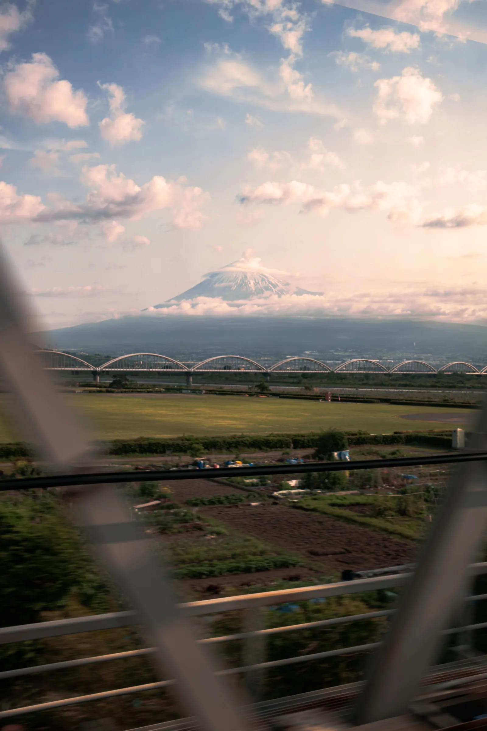 Mount Fuji under a cloud cap behind an arched bridge, seen from a train window